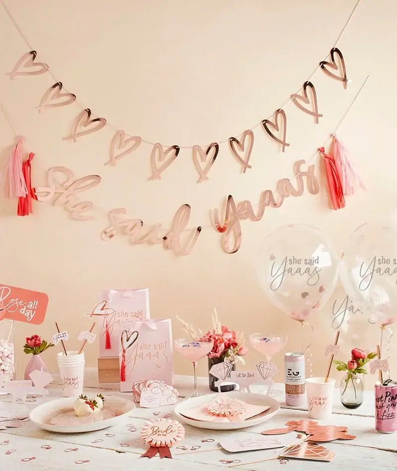 Rose gold bridal shower table setup with heart garlands, balloons, party decorations, tableware, and floral accents in soft pink tones. Rose gold bridal shower table setup with heart garlands, balloons, party decorations, tableware, and floral accents in soft pink tones.